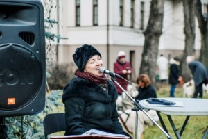 Sieviete dzied pie mikrofona, fonā redzami cilvēki un ainava. Rīgas 1. slimnīcas pasākums.