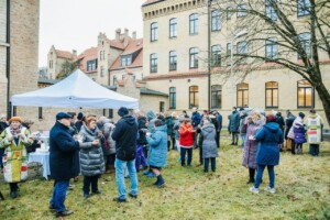 Pasākums Rīgas 1. slimnīcas pagalmā, cilvēki pulcējas zem balta teltī, baudot ēdienu un dzērienus.