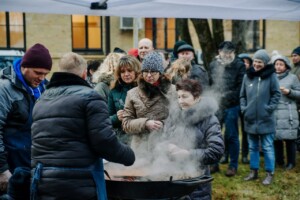 Cilvēki pulcējušies ap lielu katlu, kur tiek gatavota ēdiena porcija. Rīgas 1. slimnīcas pasākums.
