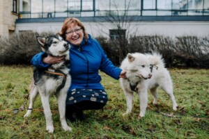 Sieviete ar sarkaniem matiem, zila jaka, pozē ar diviem suņiem, viens ir melns, otrs balts, zālē pie Rīgas 1.