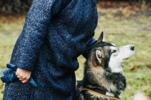 Sieviete zilā apģērbā stāv blakus husky sunim, smaidot un turot suni pie pavadas.