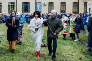 Divi cilvēki, tērpti svinīgās drēbēs, soļo pa zāli, apkārt skatās cilvēku pūlis. Rīgas 1. slimnīca fons.