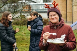 Trīs sievietes ārā, viena ar raga cepuri, tur šķīvi ar ēdienu un dakšiņu. Divas dzer kafiju no papīra krūzēm.