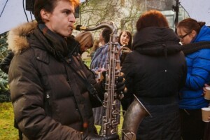 Vīrietis spēlē saksofonu, apkārt cilvēki, kas klausās un sarunājas. Notikums ārā, Rīgas 1. slimnīcas pasākums.