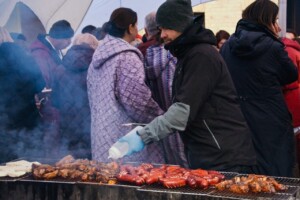 Vīrietis grilē gaļu un desas, apkārt cilvēki, dūmi no grila. Rīgas 1. slimnīca pasākums.