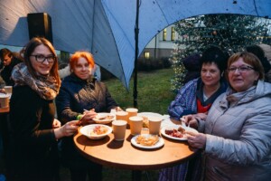 Četras sievietes stāv pie galda ar ēdienu un dzērieniem, svinot pasākumu Rīgas 1. slimnīcā.