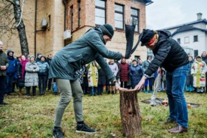 Divi cilvēki sit koku, apkārt stāv cilvēku pūlis. Rīgas 1. slimnīcas pasākums brīvā dabā.