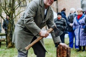 Vīrietis ar cirvi gatavojas cirst malku, apkārt stāv cilvēki. Fons: Rīgas 1. slimnīcas teritorija.