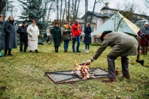 Vīrietis pie ugunskura pievieno malku, apkārt stāv cilvēki. Fons ar kokiem un telti.