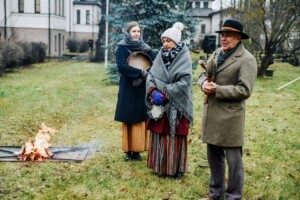 Trīs cilvēki stāv pie ugunskura, tērpti tradicionālās drēbēs, svinot pasākumu Rīgas 1. slimnīcas teritorijā.