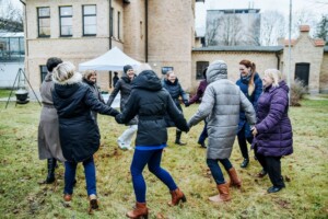 Grupas cilvēku, tērpušies siltās jakās, dejo un turas rokās Rīgas 1. slimnīcas pagalmā.