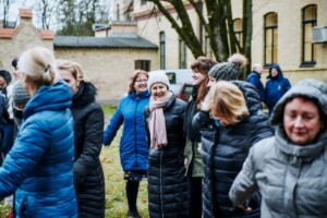 Grupa cilvēku, ģērbušies siltos mēteļos, izklaidējas ārā pie Rīgas 1. slimnīcas.