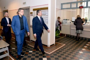rd_1slimnica_20160418 Vīrieši, ģērbušies uzvalkos, ieiet Rīgas 1. slimnīcas reģistratūrā. Redzama reģistrācijas informācija.