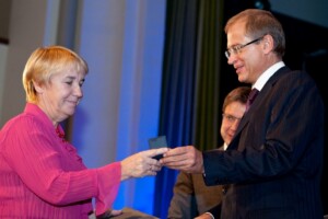 Divi cilvēki apmainās ar balvu Rīgas 1. slimnīcas pasākumā, fonā redzami dekoratīvi elementi.