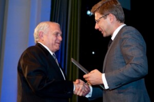 Divi vīrieši sveicinās un apmainās ar balvu Rīgas 1. slimnīcas pasākumā.