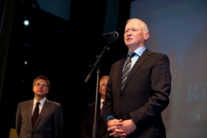 Vīrietis uz skatuves runā pie mikrofona, fonā redzams teksts "Rīgas 1. slimnīca". Divi vīrieši stāv blakus.
