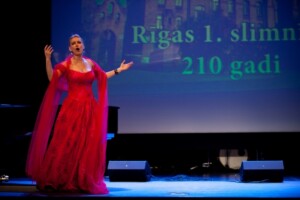 Sieviete sarkanā kleitā dzied uz skatuves, fonā teksts "Rīgas 1. slimnīca 210 gadi".