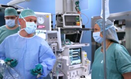Trīs mediķi operāciju telpā, valkājot maskas un ķirurģiskās drēbes, strādā pie medicīnas iekārtām.