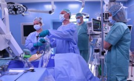 Četri medicīnas darbinieki operāciju zālē, veicot procedūru ar medicīnisko aprīkojumu.