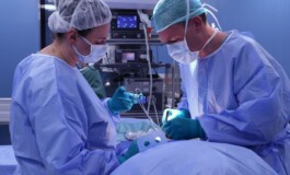 Divi ķirurgi veic operāciju, tērpti zilos medicīniskos tērpos un maskās, operācijas telpā.