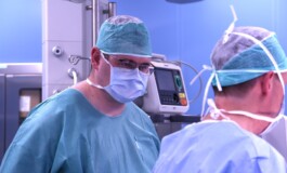 Divi ķirurgi operāciju zālē, valkājot maskas un ķirurģiskos apģērbus. Fons ar medicīnas iekārtām.