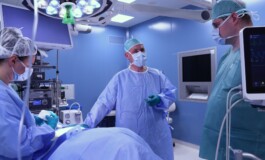 Trīs mediķi operāciju zālē, valkājot aizsargapģērbu un maskas, gatavojas veikt procedūru.