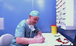 Ārsts raksta piezīmes pie galda, fona zilā krāsā, blakus medicīnas materiāliem un atkritumu tvertnei.