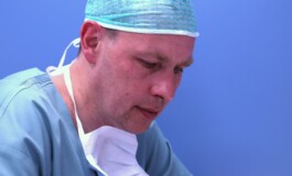 Ārsts ar operāciju tērpu un masku, koncentrējoties uz darbu Rīgas 1. slimnīcā. Fons zils.